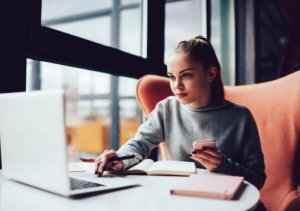 girl sitting in wing back chair with a diary in front and laptop on desk while holding mobile phone