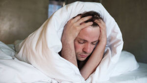 man laying on bed holding head with doona over them